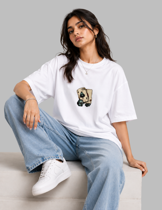 A woman wearing a white oversized graphic t-shirt with a playing card design, paired with light blue wide-leg jeans and white sneakers, sitting on a light gray platform against a solid gray background.