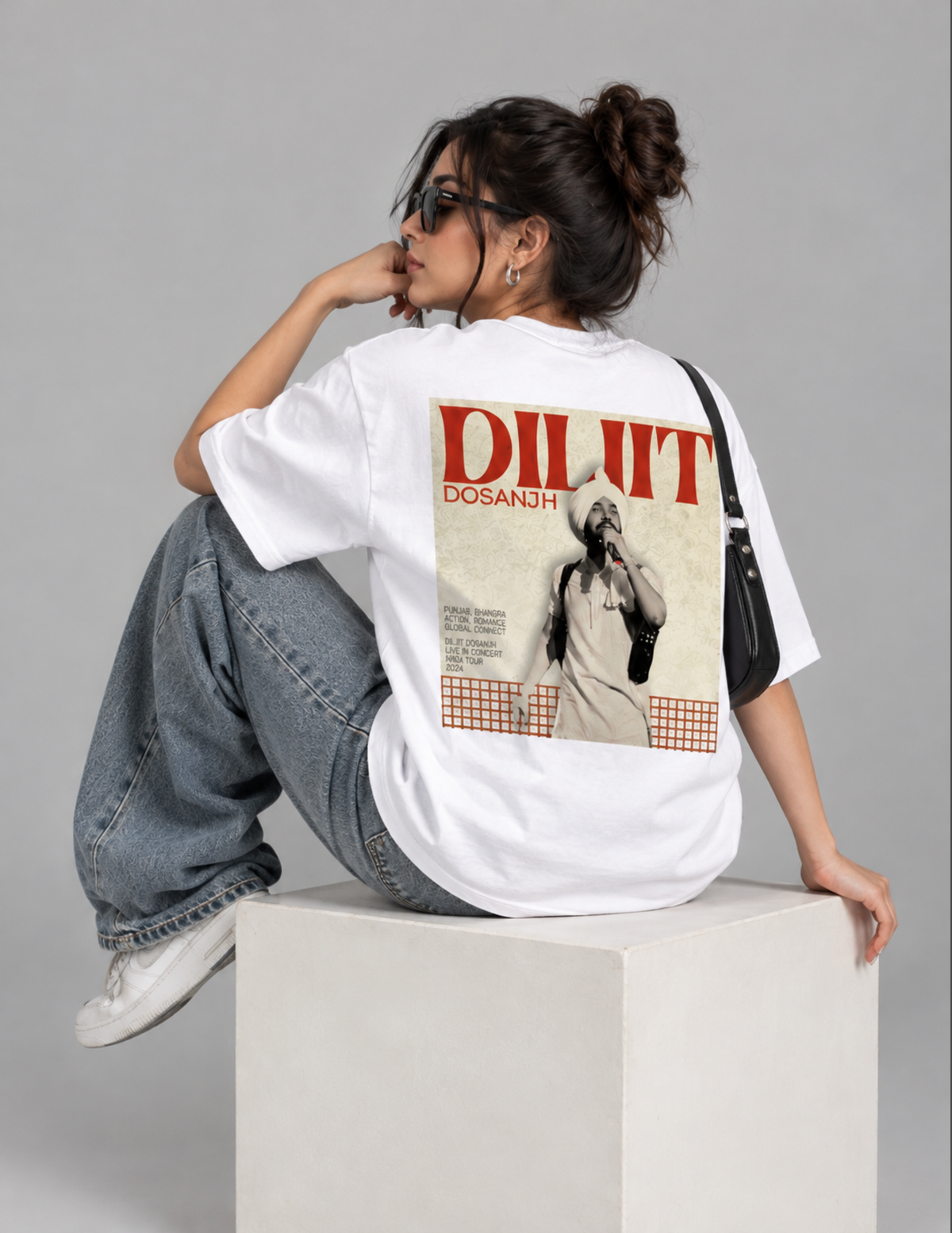 A woman wearing a black oversized t-shirt with a large graphic print on the back, sitting on a concrete block against a dark gray studio background.