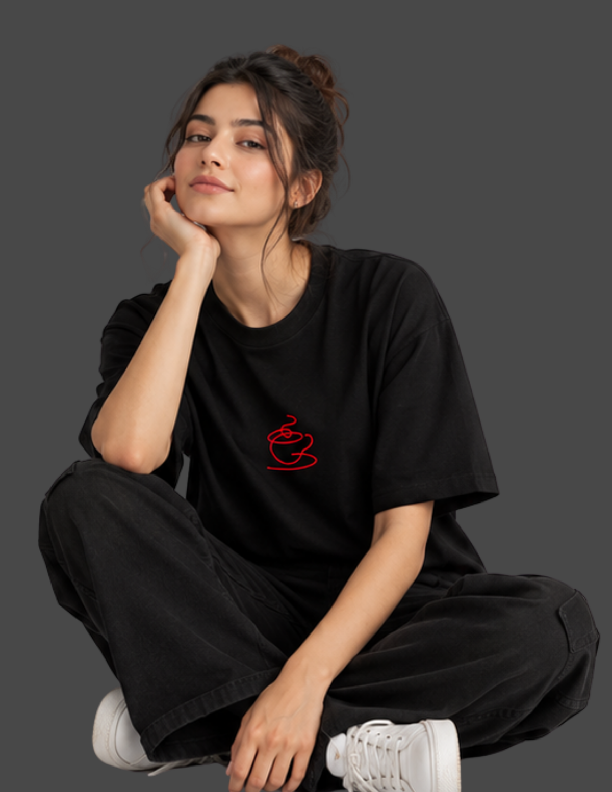 A woman sitting cross-legged against a dark gray background, wearing a black oversized t-shirt with a small red coffee cup graphic on the left chest, paired with black sweatpants and white sneakers.