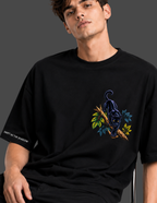 A man wearing a black oversized t-shirt with a graphic of a black panther on a branch with green and blue leaves on the chest, and white text on the sleeve reading 'SHIRT AS THE SHADOW'.