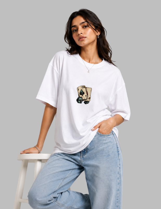 A woman wearing an oversized white t-shirt with a graphic of a playing card on the chest, paired with light blue jeans, leaning on a white stool against a solid gray background.