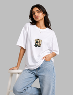 A woman wearing an oversized white t-shirt with a graphic of a playing card on the chest, paired with light blue jeans, leaning on a white stool against a solid gray background.