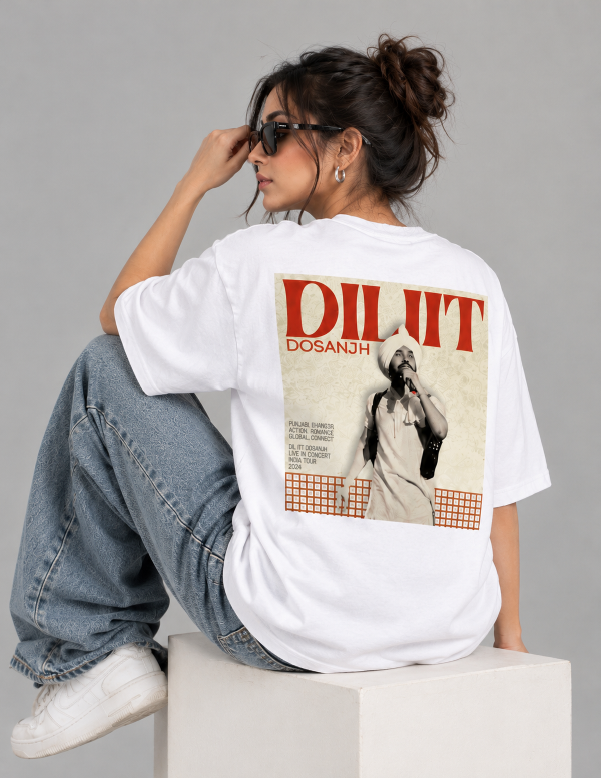 A woman wearing a black oversized t-shirt with a large graphic print on the back, sitting on a concrete block against a dark gray studio background.