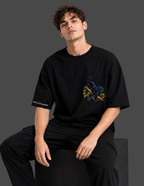 A man wearing a black short-sleeve t-shirt with a colorful panther graphic on the left chest and white text on the right sleeve, paired with black pants, seated against a dark gray studio background.
