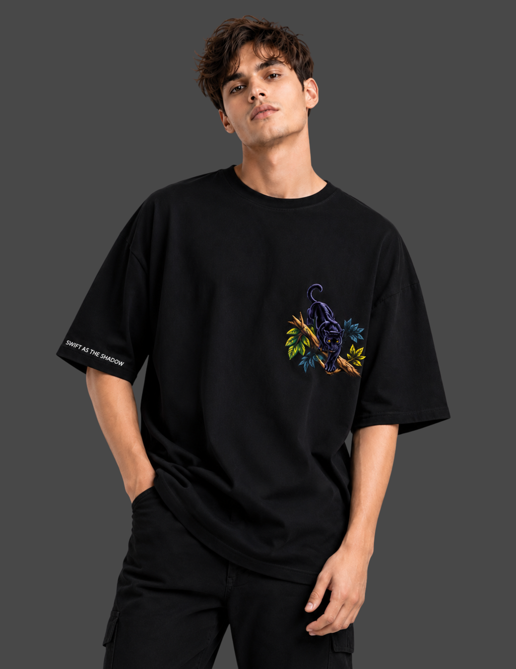 A man wearing a black oversized t-shirt with a colorful graphic of a panther and leaves on the left chest, paired with black pants, set against a dark gray studio background.