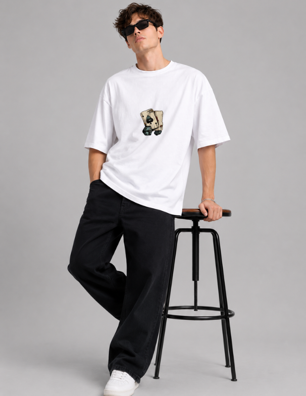 A man wearing a white oversized T-shirt with a small graphic print on the chest, paired with black pants and white sneakers, leaning against a black stool on a gray studio background.