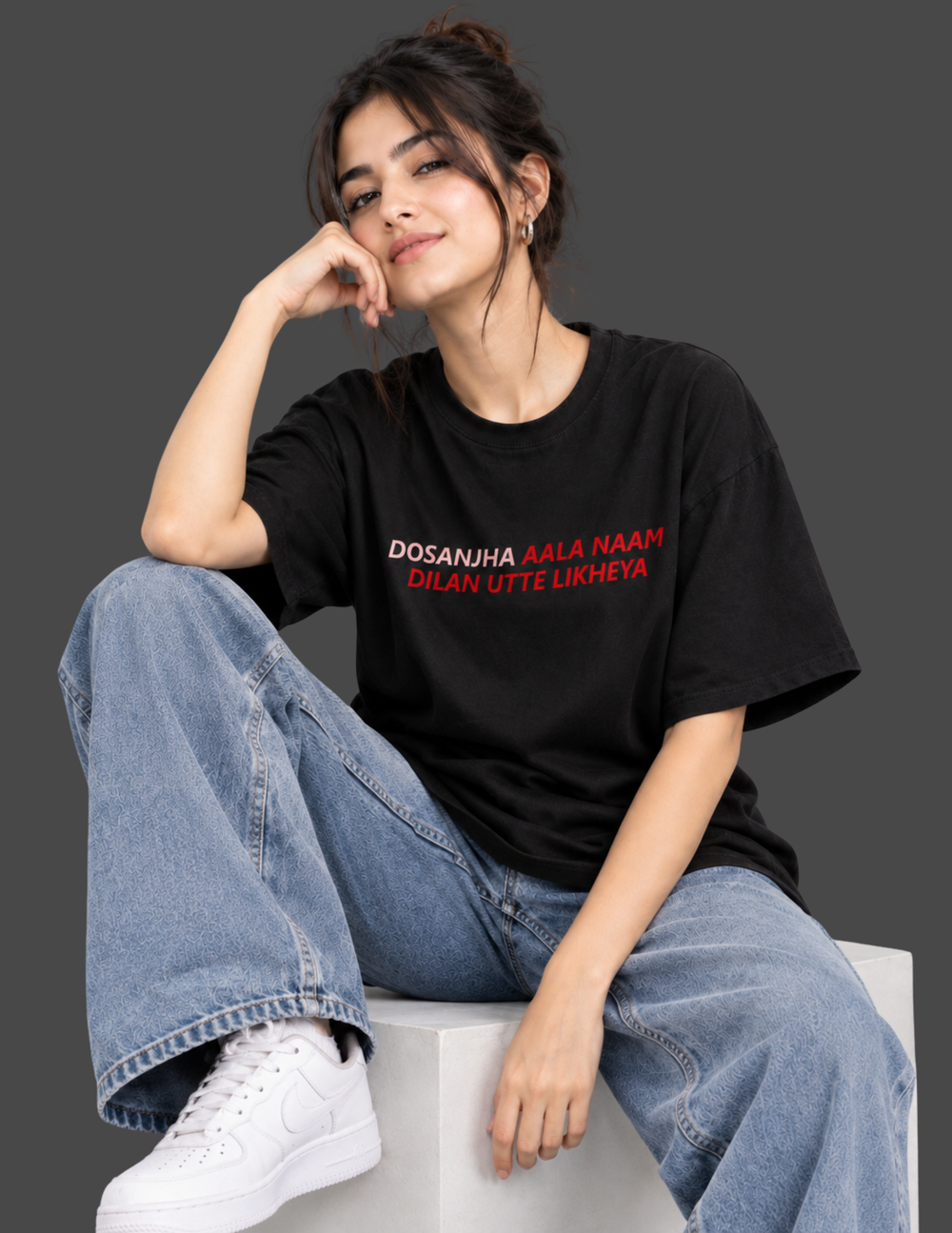 A woman sits on a white block wearing a black graphic t-shirt with red and white text, paired with light blue wide-leg jeans and white sneakers, against a dark gray studio background.
