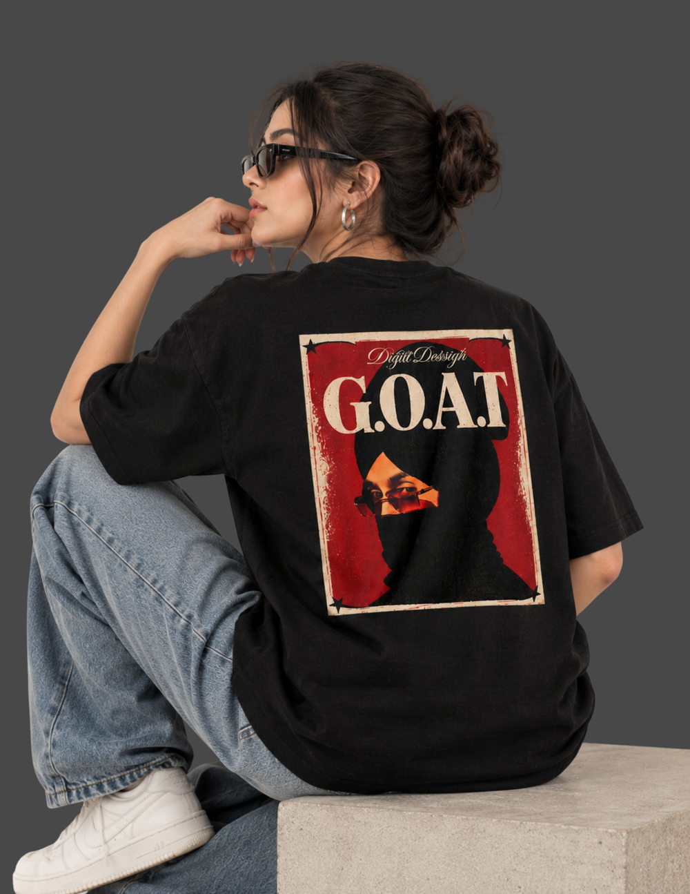 A woman wearing a black oversized t-shirt with a large graphic print on the back, sitting on a concrete block against a dark gray studio background.
