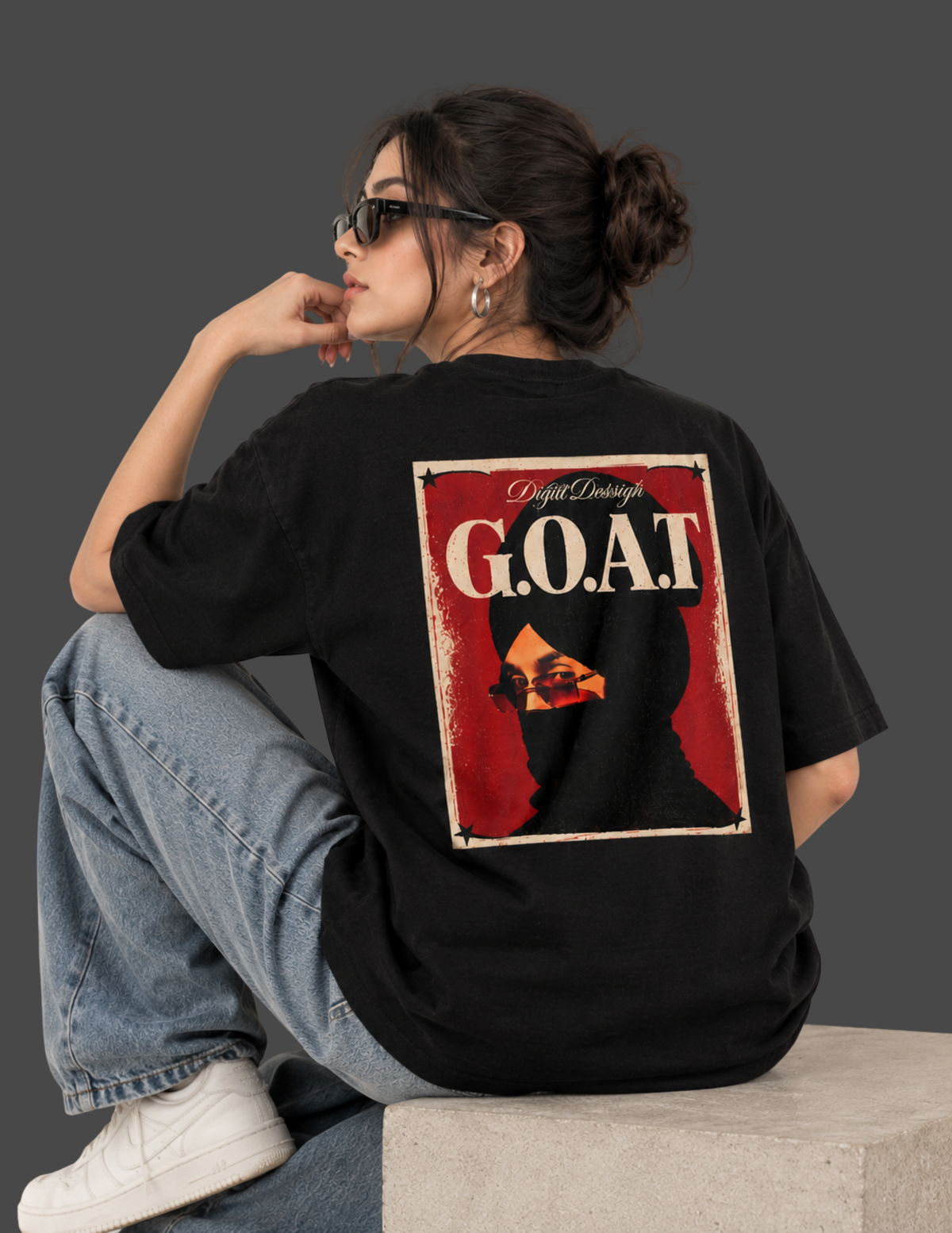 A woman wearing a black oversized t-shirt with a large graphic print on the back, sitting on a concrete block against a dark gray studio background.