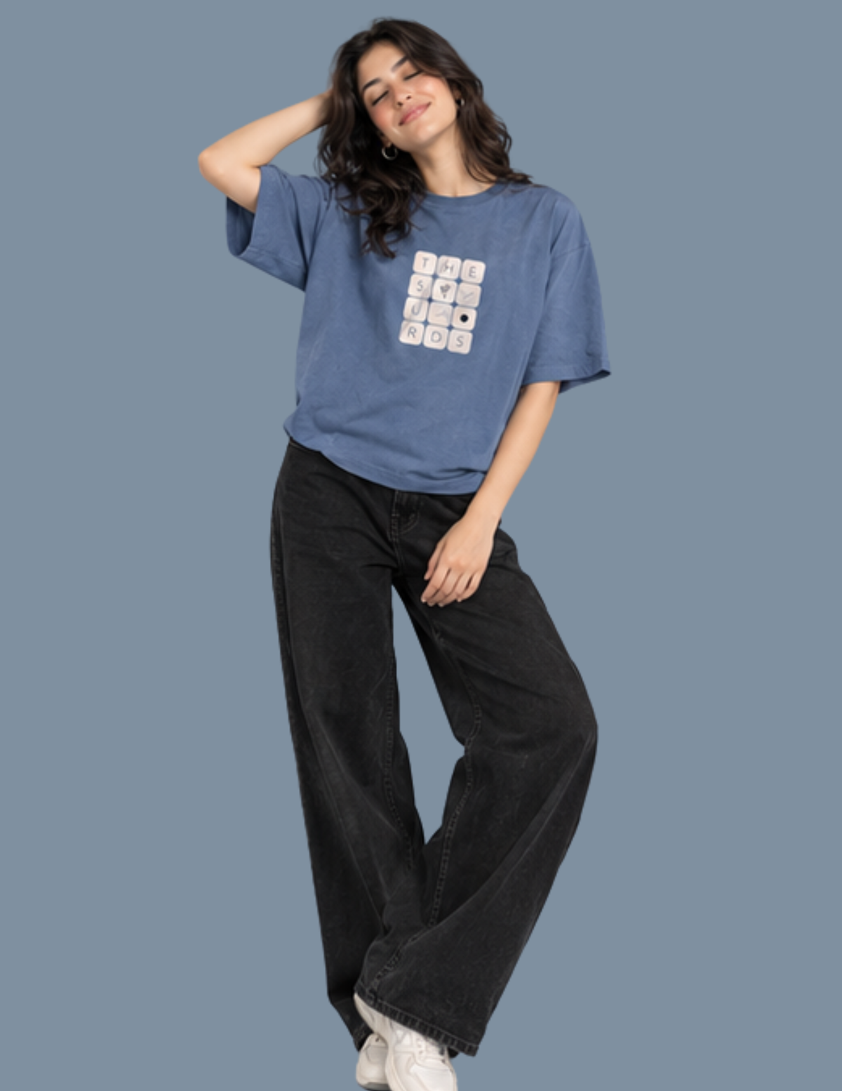 A woman modeling a blue short-sleeve graphic t-shirt with a white and black grid design on the front, paired with black wide-leg jeans and white sneakers, set against a solid slate blue background.