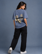 A woman wearing a dusty blue t-shirt with a large graphic print on the back, paired with black cargo pants and white sneakers, standing against a solid slate blue background.