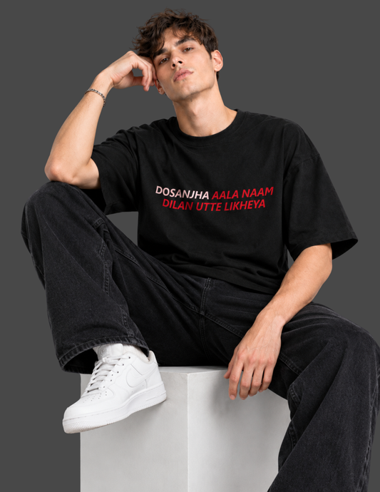 A man wearing a black graphic t-shirt with red and white text, paired with black jeans and white sneakers, sitting on a white block against a dark gray background.