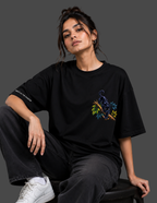 A woman wearing a black oversized graphic T-shirt with a colorful panther and floral design on the chest, paired with dark jeans and white sneakers, seated against a dark gray background.