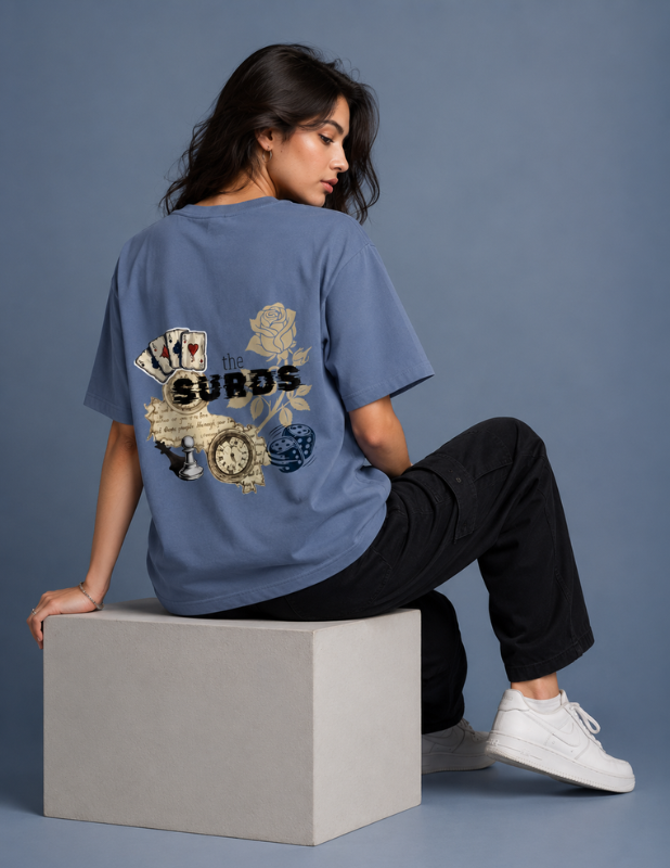 A woman sits on a gray block wearing a blue graphic t-shirt with a vintage-style design featuring playing cards, a rose, and a pocket watch, paired with black pants and white sneakers against a blue-gray studio background.