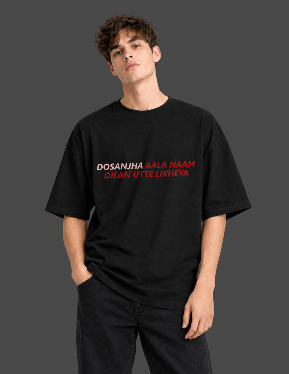 A man wearing a black oversized t-shirt with the text 'DOSANJA AALA NAAM DILAN UTTE LIKHEYA' printed in white and red on the chest, posed against a dark gray studio background.