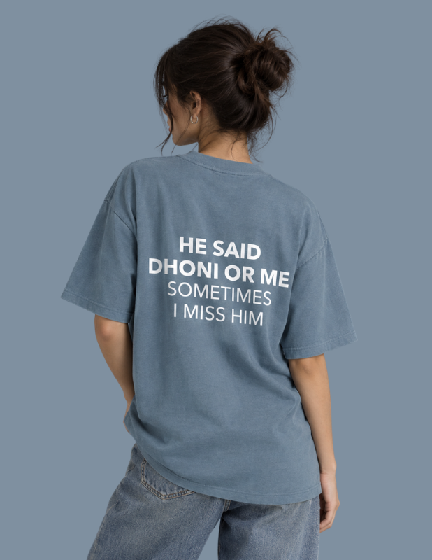 A woman shown from behind wearing an oversized dusty blue graphic T-shirt with white text that reads 'HE SAID DHONI OR ME SOMETIMES I MISS HIM', paired with light-wash jeans against a solid slate-blue background.