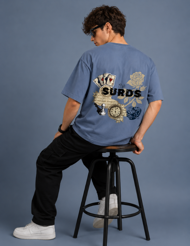 A man wearing a light blue graphic t-shirt with a large back print featuring playing cards, a rose, a clock, and the text 'the SURDS', sitting on a black metal stool against a solid blue-gray studio background.