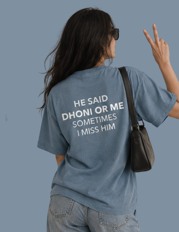 A woman shown from behind wearing a faded blue graphic t-shirt with the text 'HE SAID DHONI OR ME SOMETIMES I MISS HIM' printed in white. She is wearing light-wash jeans, sunglasses, and carrying a black shoulder bag against a solid blue-gray background.