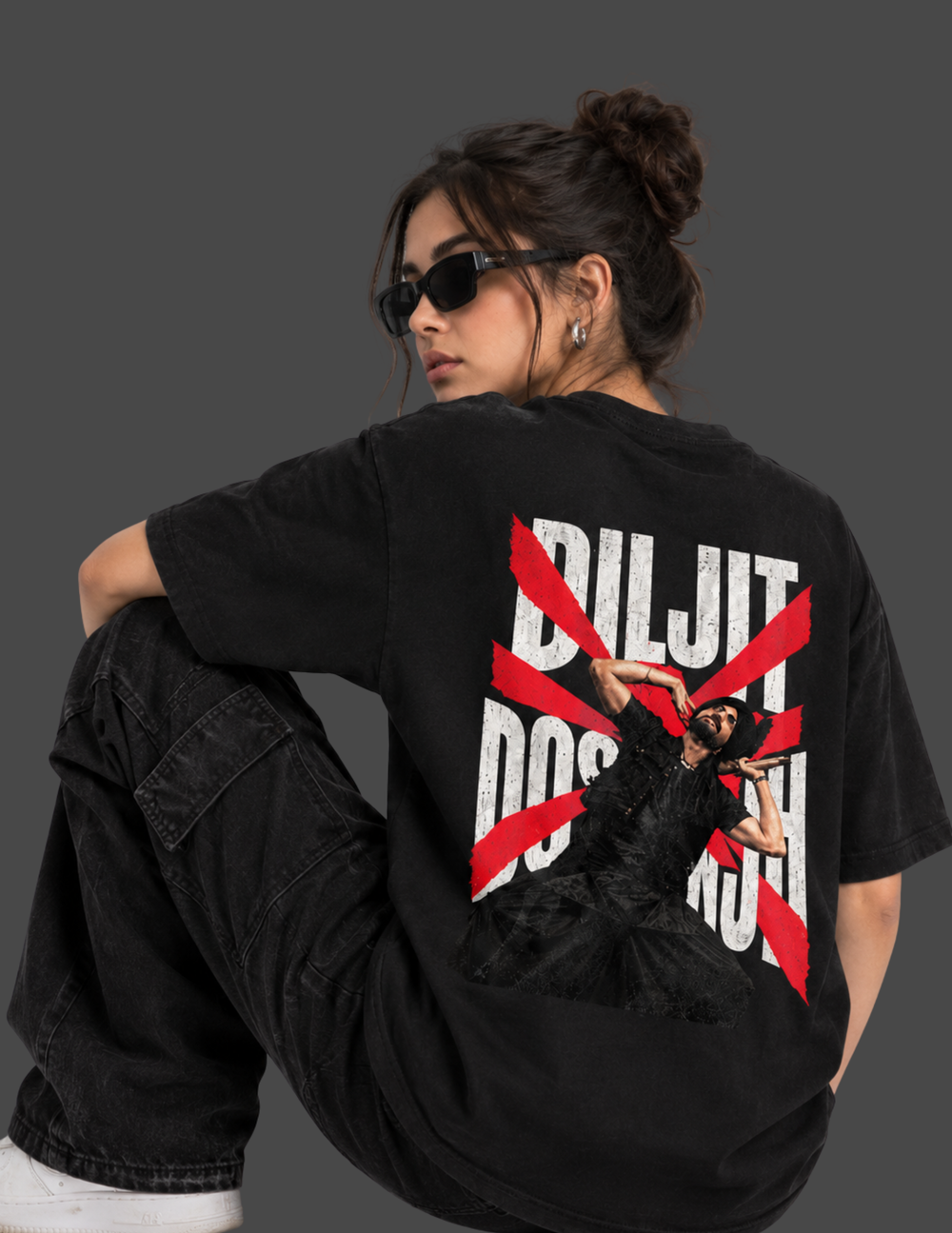 A woman wearing a black oversized t-shirt with a large graphic print on the back, sitting on a concrete block against a dark gray studio background.
