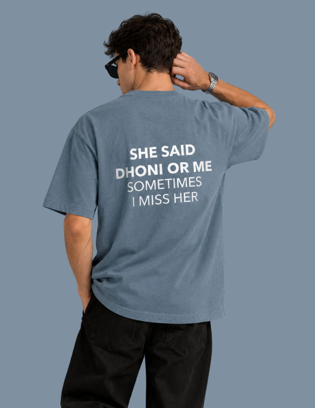A man shown from behind wearing a faded blue t-shirt with white printed text that reads 'SHE SAID DHONI OR ME SOMETIMES I MISS HER'.