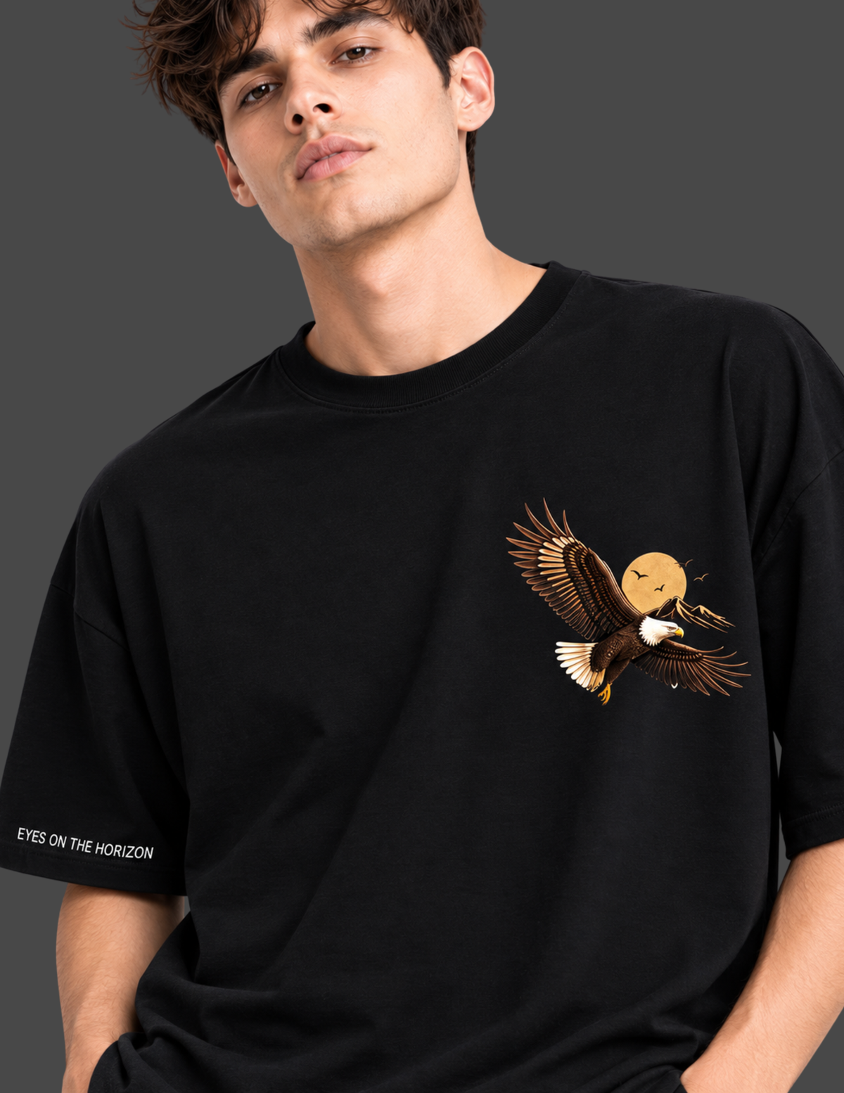 A man wearing a black oversized t-shirt with a graphic of a flying bald eagle holding a moon and a fish on the left chest, with white text on the right sleeve reading 'EYES ON THE HORIZON'.