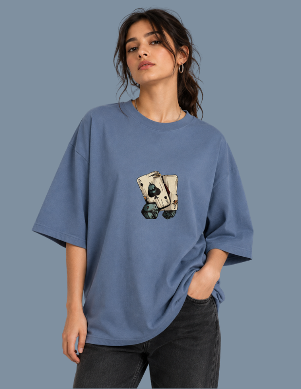 A woman wearing an oversized dusty blue t-shirt with a graphic of two playing cards on the front, paired with dark gray jeans, set against a solid slate blue background.
