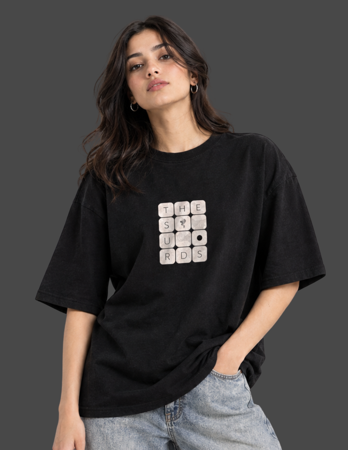 A woman wearing a black oversized t-shirt with a white crossword puzzle graphic on the front that spells out 'THE STUNROS', paired with light-wash jeans, set against a dark gray studio background.