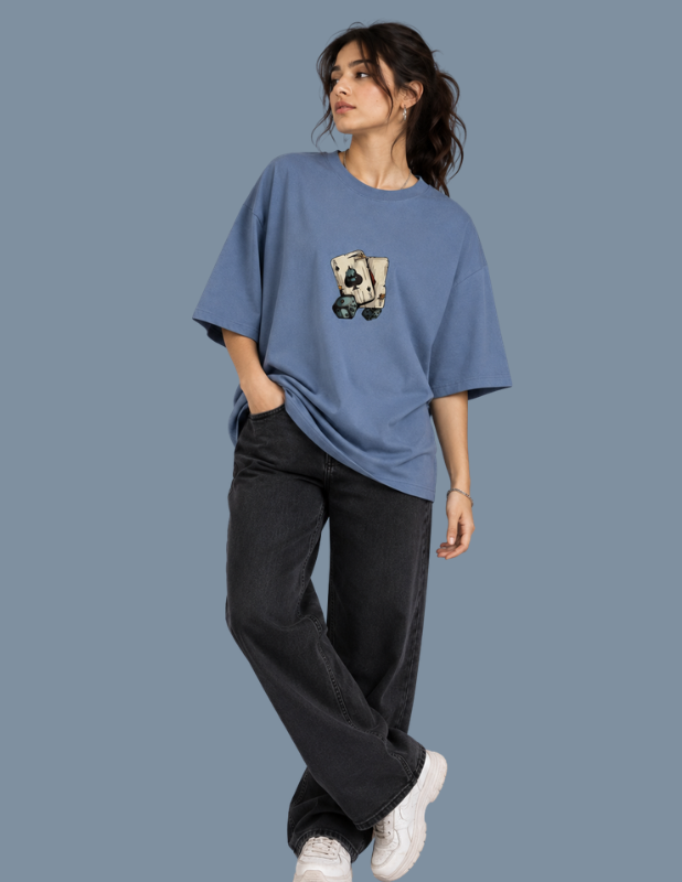 A woman modeling an oversized dusty blue t-shirt with a graphic of a hand holding a card on the front, paired with dark gray wide-leg jeans and white sneakers, set against a solid slate blue background.