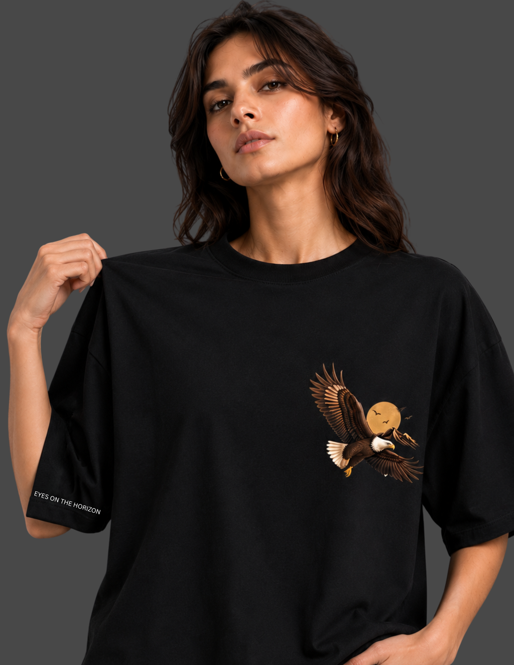 A woman wearing an oversized black t-shirt with a graphic of a bald eagle carrying a smiling sun on its wing. The t-shirt has a small white text print on the left sleeve that reads 'EYE ON THE PREDATOR'.