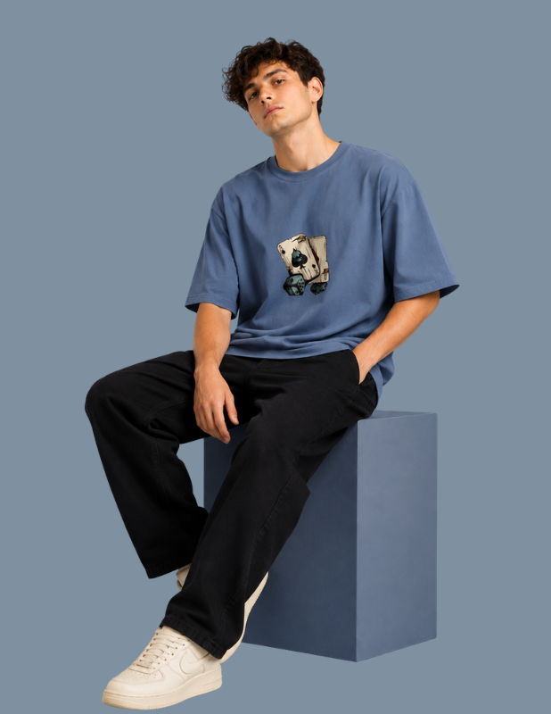 A young man wearing a blue crewneck t-shirt with a graphic print of a cartoon character on a skateboard, paired with black pants and white sneakers, sitting on a blue block against a solid blue-gray background.