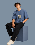 A young man wearing a blue crewneck t-shirt with a graphic print of a cartoon character on a skateboard, paired with black pants and white sneakers, sitting on a blue block against a solid blue-gray background.