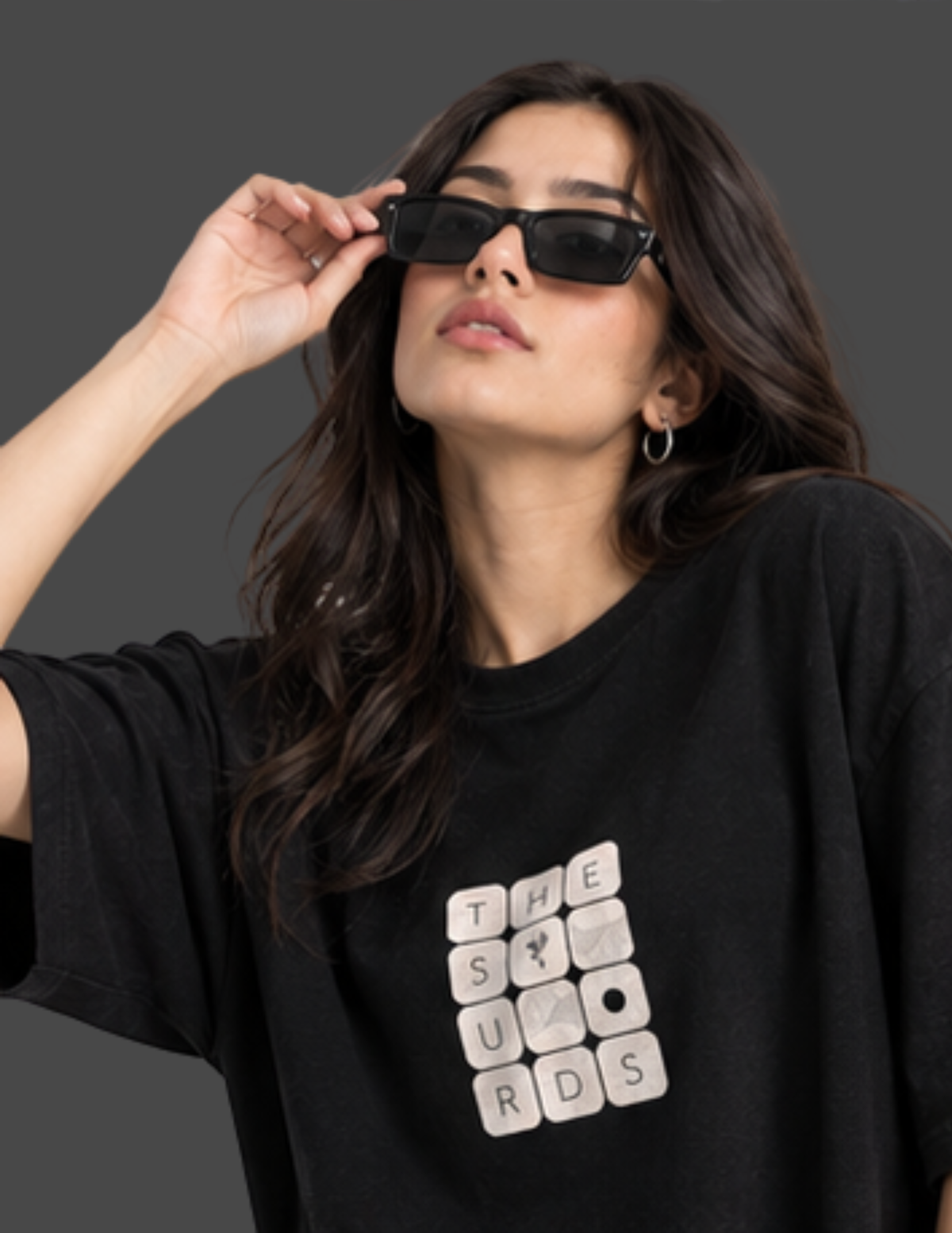 A woman wearing a black oversized t-shirt with a white Scrabble-style letter grid graphic on the front, paired with black sunglasses, posed against a dark gray studio background.