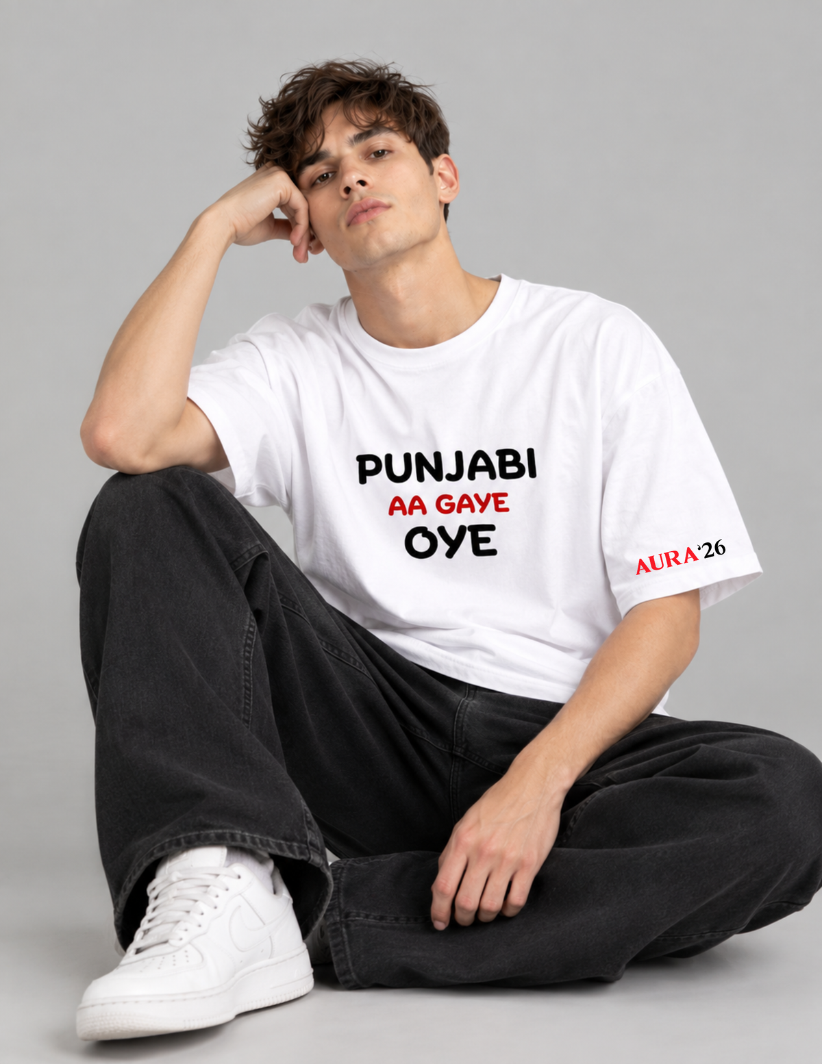 A man sitting on the floor wearing a white graphic t-shirt with the text 'PUNJABI AA GAVE OYE' in black and red, paired with black jeans and white sneakers, against a solid gray studio background.