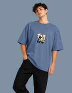 A man wearing a blue oversized t-shirt with a graphic of playing cards on the chest, paired with black pants, set against a solid blue-gray background.