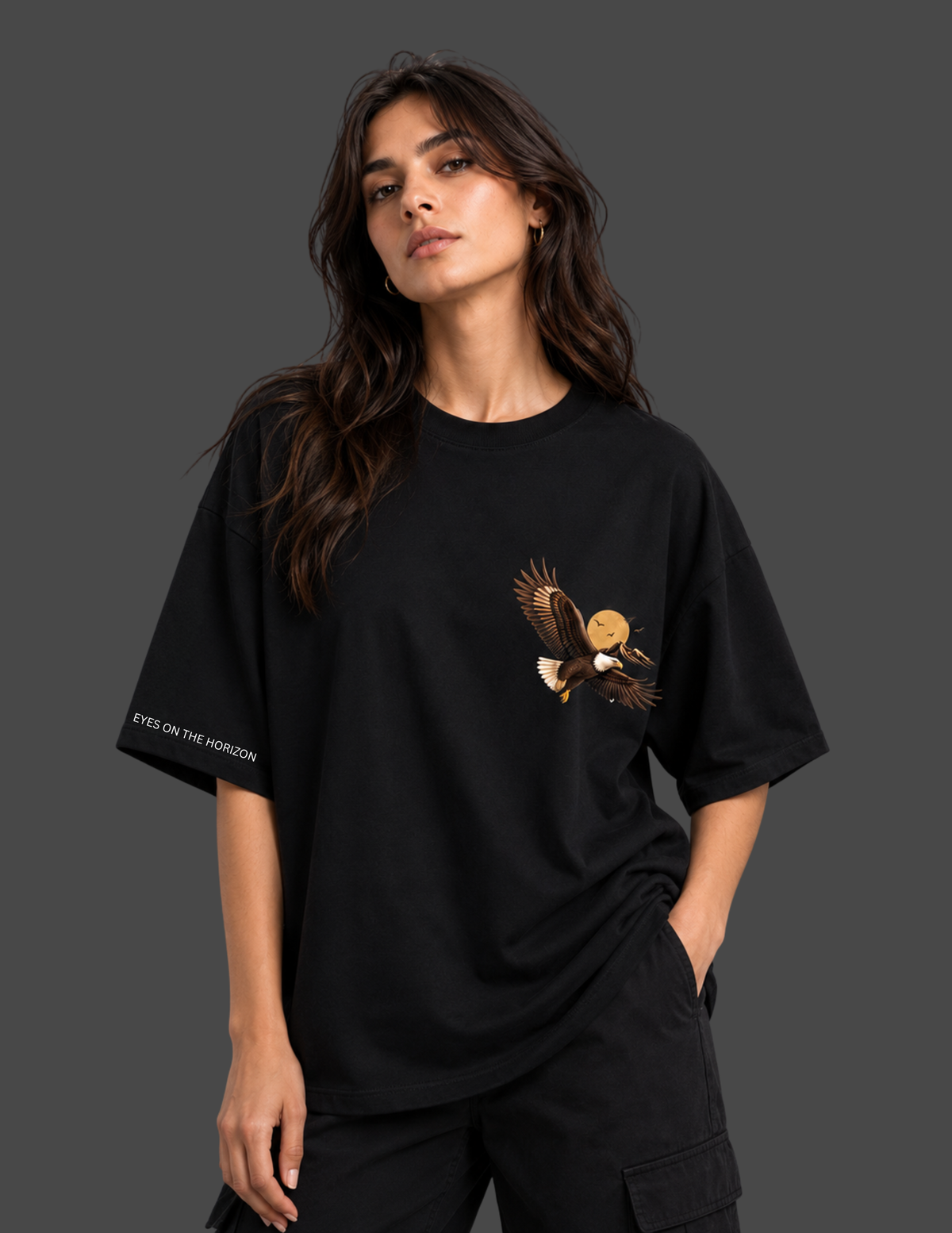 A woman modeling a black oversized t-shirt with a detailed eagle graphic on the left chest and white text on the sleeve, set against a solid dark gray background.