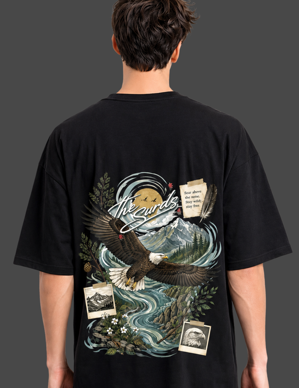 A man viewed from behind wearing a black t-shirt with a large, detailed graphic print on the back. The graphic features an eagle flying over a mountain and river landscape with vintage-style postcards and the text 'The Wilds'.