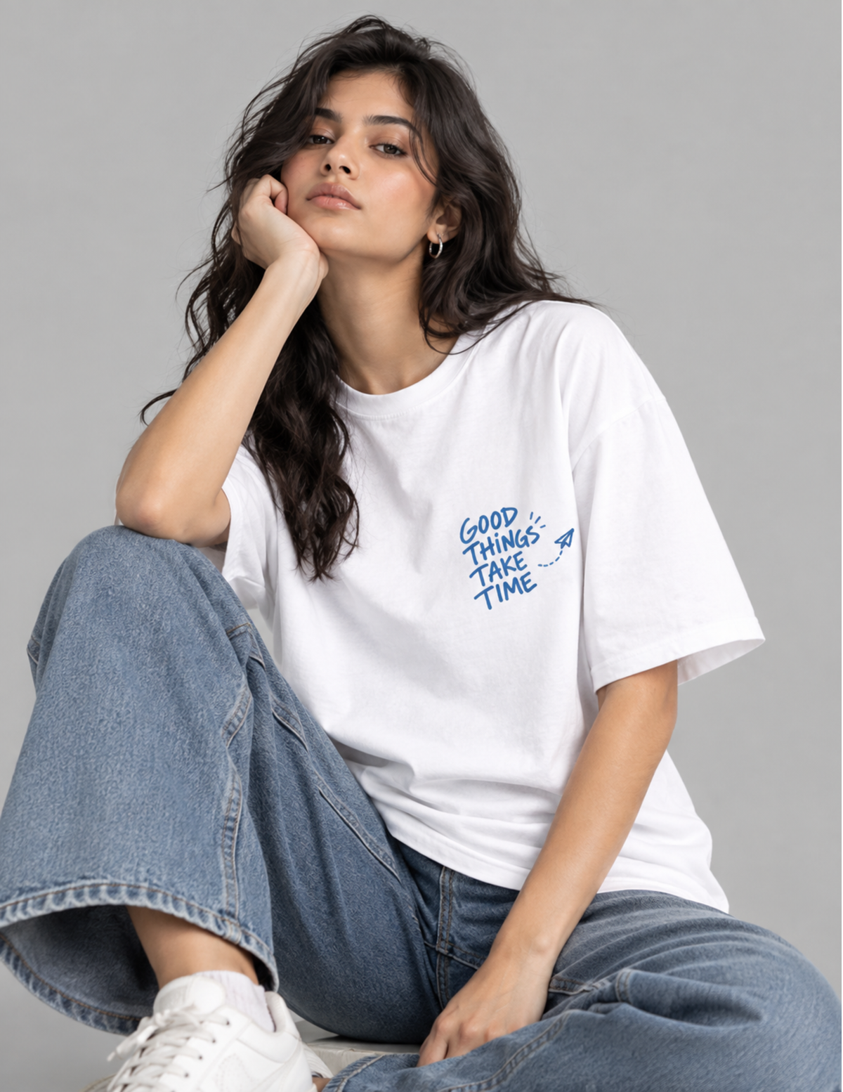 A woman sitting cross-legged wearing a white crewneck t-shirt with a blue graphic that reads 'GOOD THINGS TAKE TIME' and blue jeans, posed against a solid gray studio background.