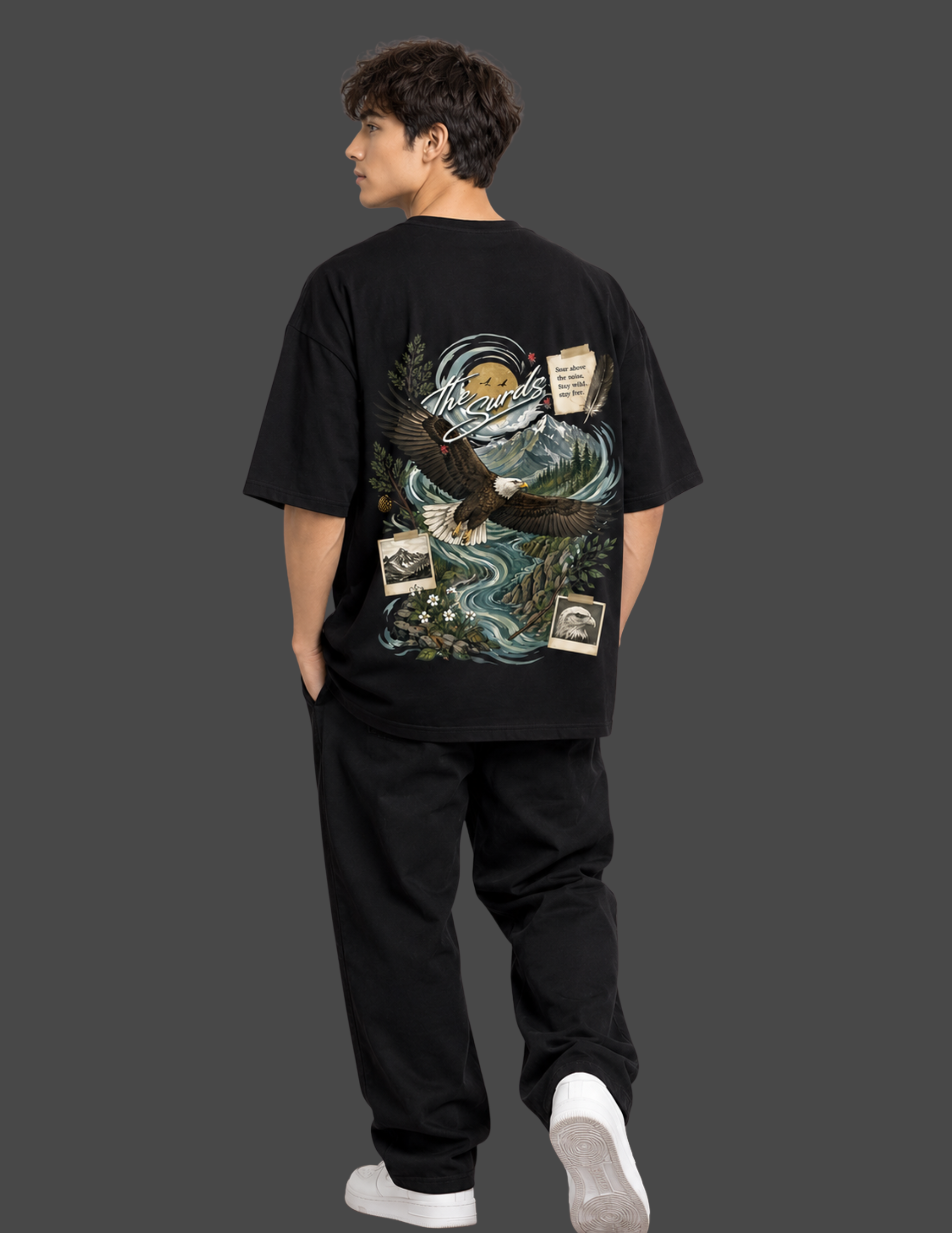 A man shown from the back wearing a black oversized graphic T-shirt with a large illustrated eagle, waves, and small card designs on the back, paired with black pants and white sneakers against a dark gray studio background.