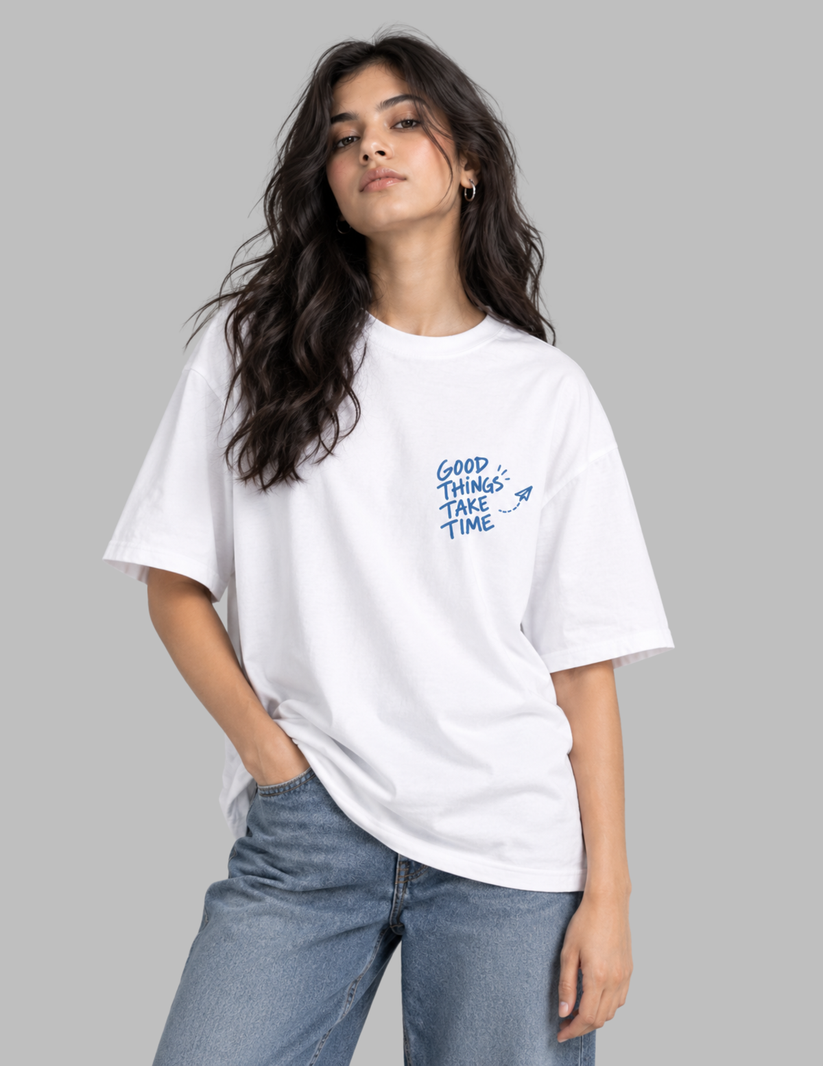 A woman wearing a white oversized t-shirt with blue text reading 'GOOD THINGS TAKE TIME' on the left chest, paired with light blue jeans, against a solid gray background.