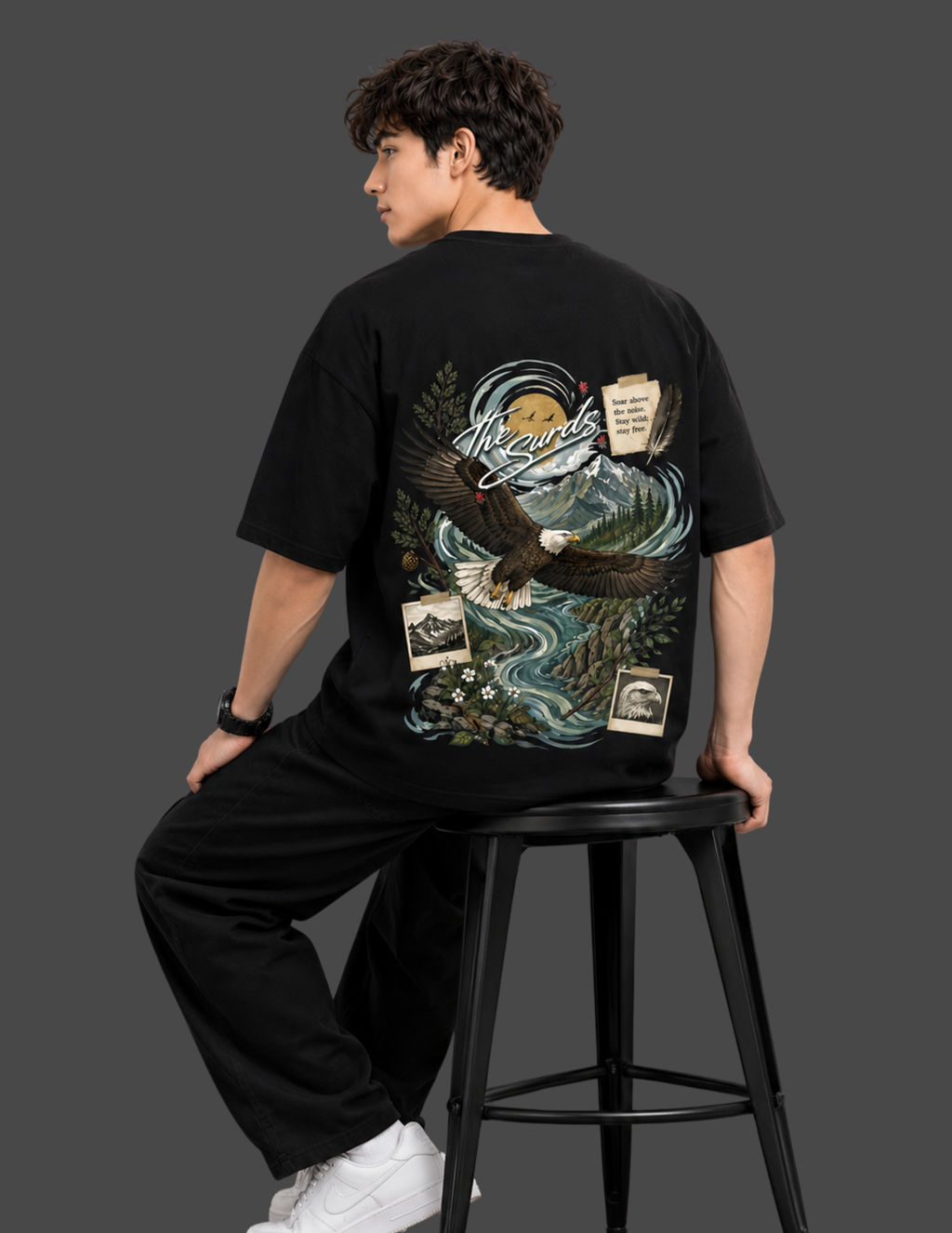 A man wearing a black graphic t-shirt and black pants, sitting on a black stool against a dark gray background. The back of the t-shirt features a large detailed graphic of an eagle over a river landscape with a moon and text.