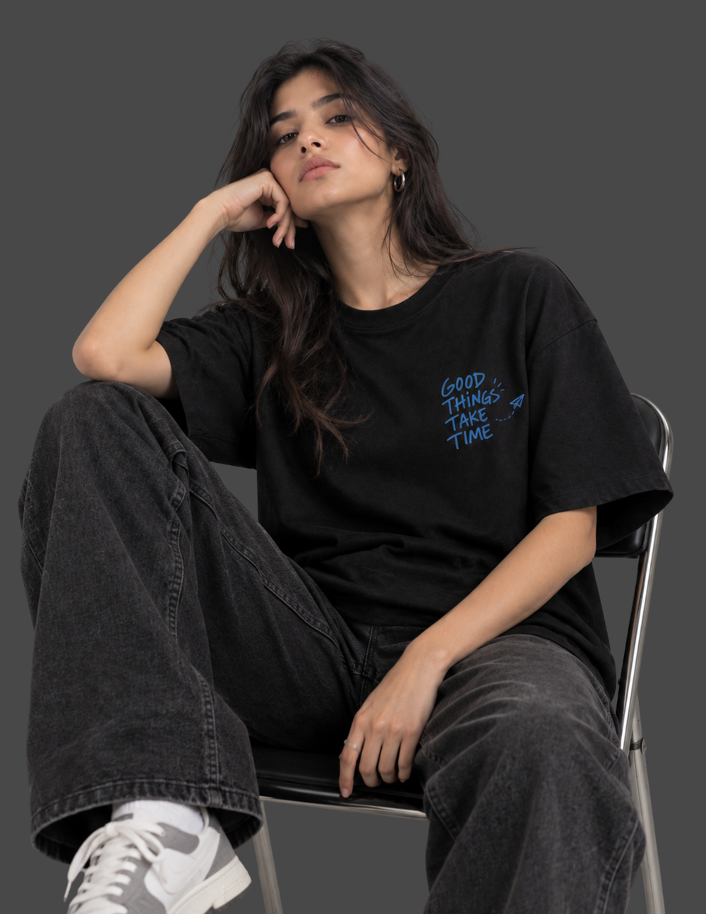 A woman sits on a metal chair wearing a black oversized graphic T-shirt with blue text reading 'GOOD THINGS TAKE TIME' on the left chest, paired with dark gray jeans and white sneakers, against a dark gray studio background.