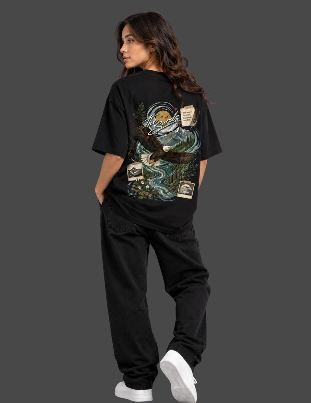 A woman modeling a black oversized graphic T-shirt from the back, paired with black sweatpants and white sneakers, against a solid dark gray studio background.