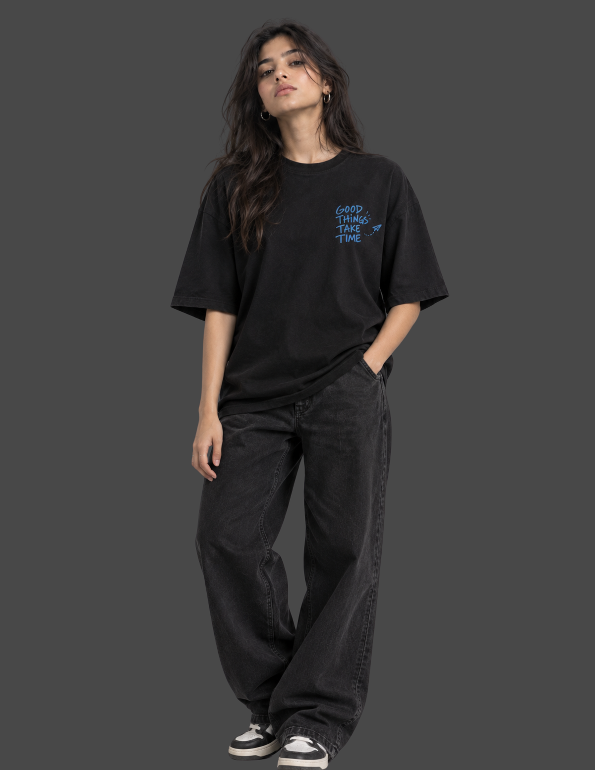 A woman modeling a black oversized t-shirt with a small blue graphic on the left chest that reads 'GRIP THE DYE TEES', paired with dark wide-leg jeans and black-and-white sneakers, against a solid dark gray background.