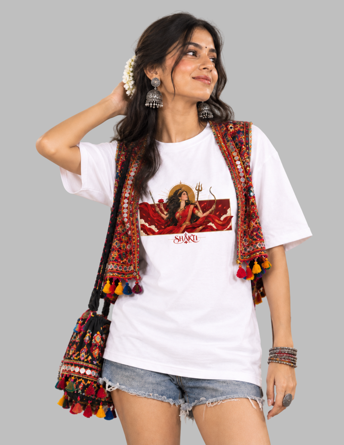A woman modeling a white graphic t-shirt featuring a red and gold illustration of the Hindu goddess Lakshmi, styled with a colorful embroidered ethnic-style vest and denim shorts against a solid gray background.
