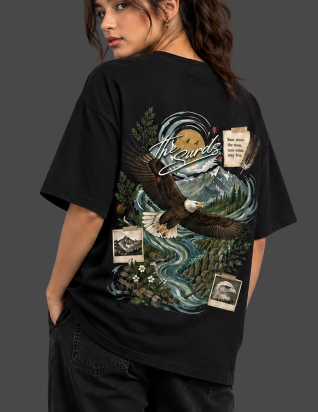 A woman shown from the back wearing a black graphic T-shirt with a large eagle, mountain, river, and text print on the back, paired with dark jeans against a dark gray studio background.