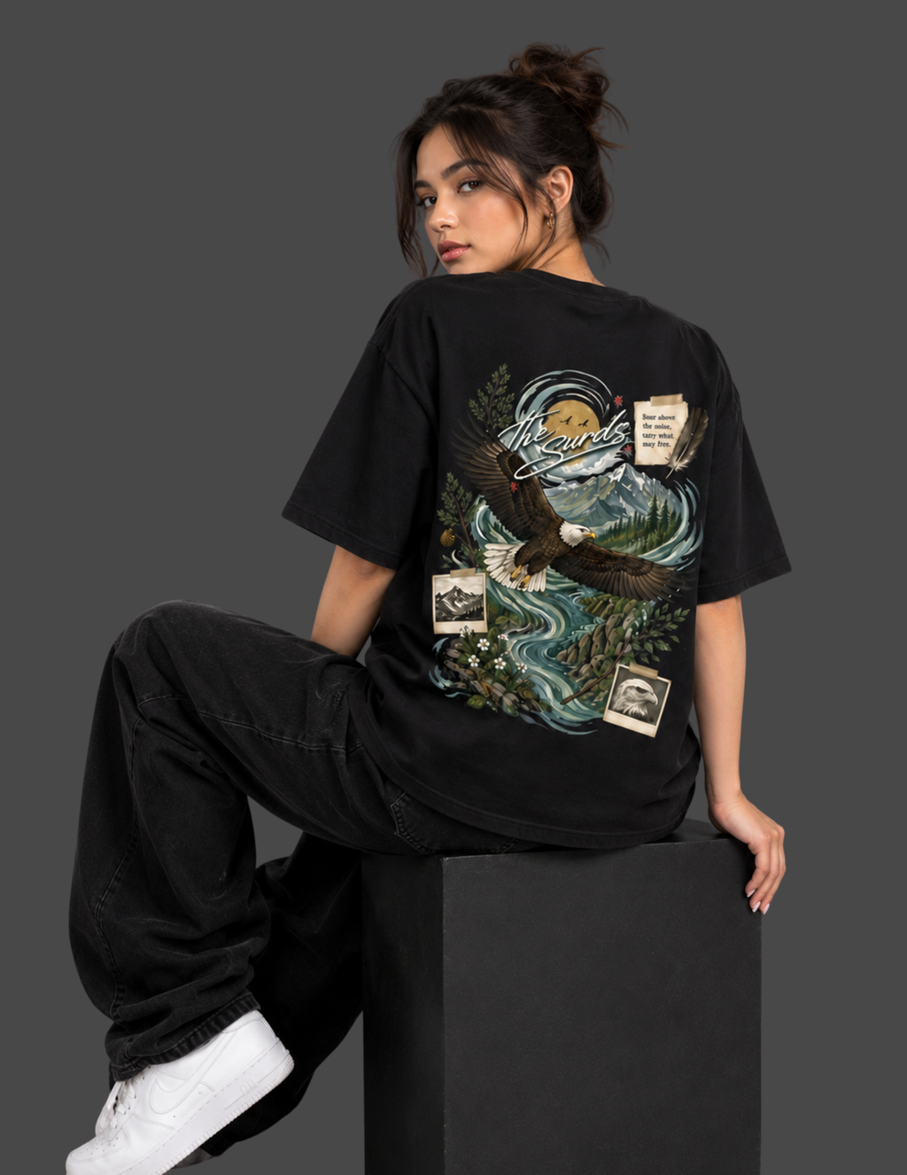 A woman wearing a black oversized graphic T-shirt and black baggy pants, sitting on a black block against a dark gray studio background. The back of the shirt features a large detailed graphic with an eagle, river, trees, and small vintage-style label patches.