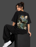 A woman wearing a black oversized graphic T-shirt and black baggy pants, sitting on a black block against a dark gray studio background. The back of the shirt features a large detailed graphic with an eagle, river, trees, and small vintage-style label patches.