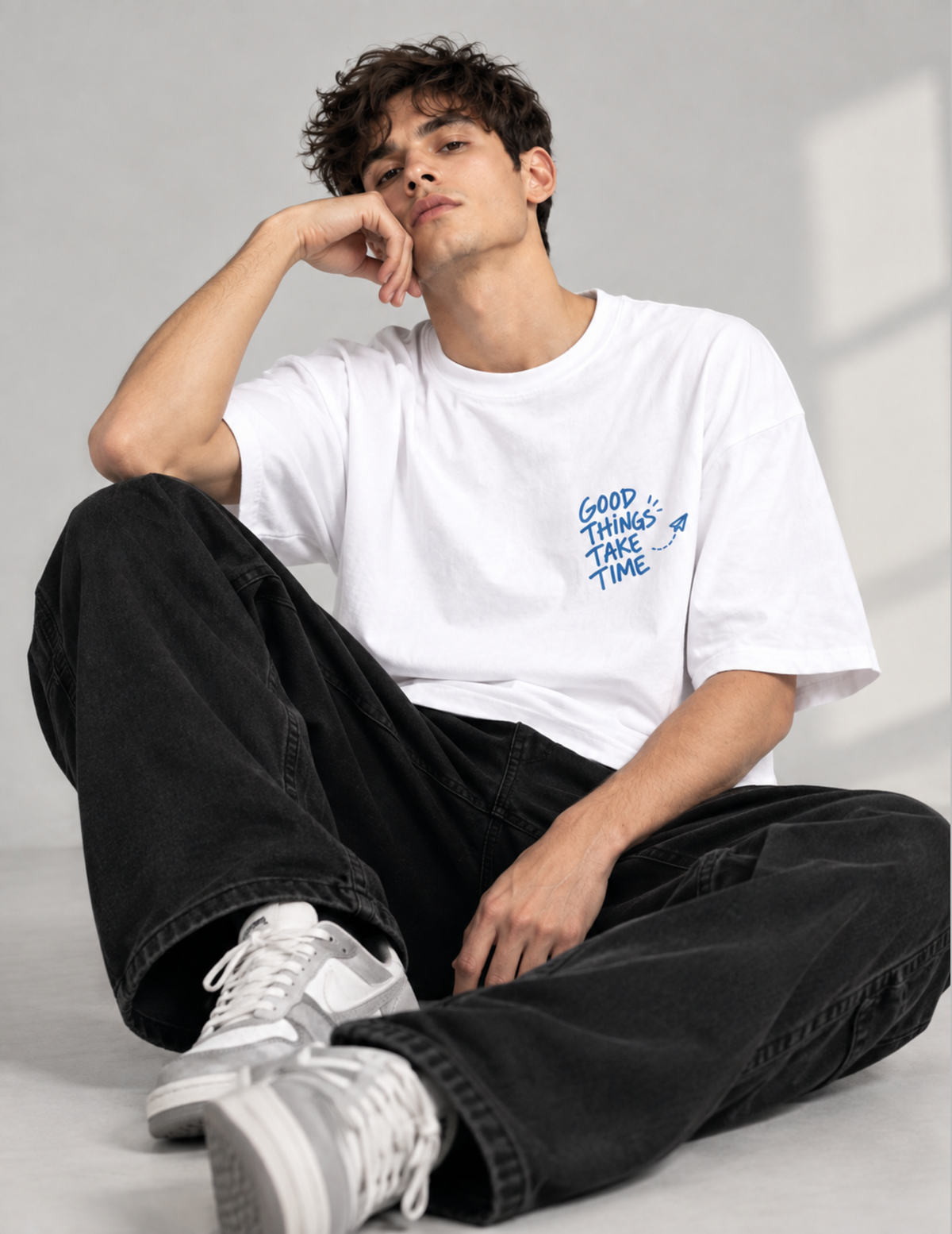 A man wearing a black t-shirt with a large graphic print on the back, sitting on a white block against a dark gray background. The graphic features a blue square with the text 'GOOD THINGS TAKE TIME' and small illustrations of people.