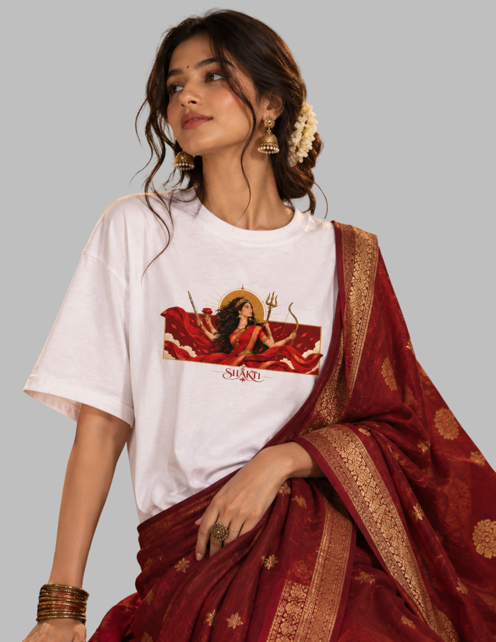A woman wearing a white graphic t-shirt with a red and gold Shiva print, paired with a deep red and gold patterned saree, set against a solid gray background.