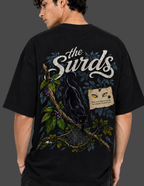 A man wearing a black graphic t-shirt, viewed from the back. The shirt features a large design with the text 'the Surds' in white script, a black cat on a tree branch with green leaves and purple berries, and a small parchment note with yellow eyes.